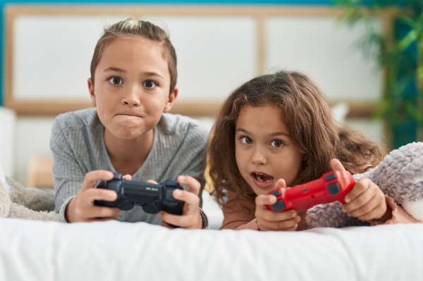Two children lying on a bed, holding PlayStation controllers in this image from Shutterstock