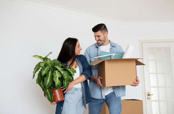 A husband is holding a box and the wife is holding a plant in this image from Shutterstock.