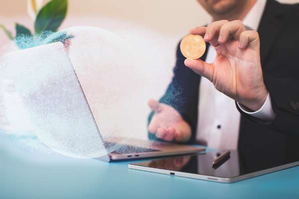 Person holding up a Bitcoin with a laptop on the desk in this image from Shutterstock.