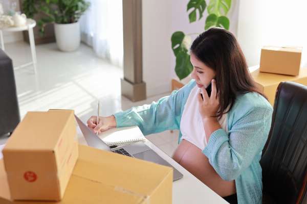 A woman sits at a desk on the phone, writing something in a notebook. A laptop is open in front of her. There are boxes throughout the room. 