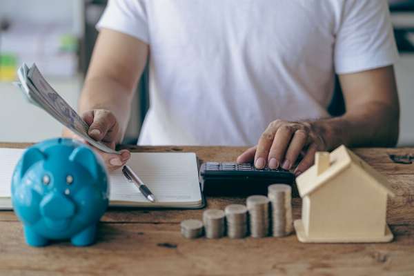 A man sits at a desk that has a notebook, calculator, cash, a wooden house, and a piggy bank in this image from Shutterstock