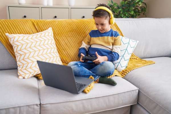 A boy in a striped shirt and headphones sits on a couch, playing a video game on a laptop
